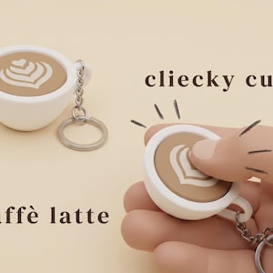 Può includere: Portachiavi a forma di tazza di caffè bianca e beige con un design a latte art. Il portachiavi ha una catena e un anello argentati. Le parole "cliecky cupz" e "caffè latte" sono visibili.