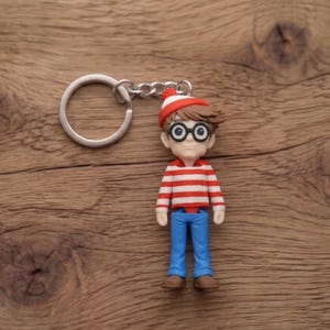 Pode incluir: Um porta-chaves com uma figura de um personagem vestindo uma camisa listrada vermelha e branca, calças azuis e um chapéu vermelho e branco. A figura tem óculos pretos e cabelo castanho. O porta-chaves é de metal.