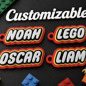 Op de afbeelding: Vier aanpasbare plastic sleutelhangers met de namen NOAH, LEGO, OSCAR en LIAM in witte blokletters op een zwarte achtergrond met gele en rode randen. Het woord Customizable staat in het wit boven de sleutelhangers.
