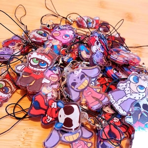 May include: A collection of colorful acrylic charms with cartoon character designs. The charms feature various figures in red, purple, and blue, each with a black string for attachment. The background is a light wood surface.