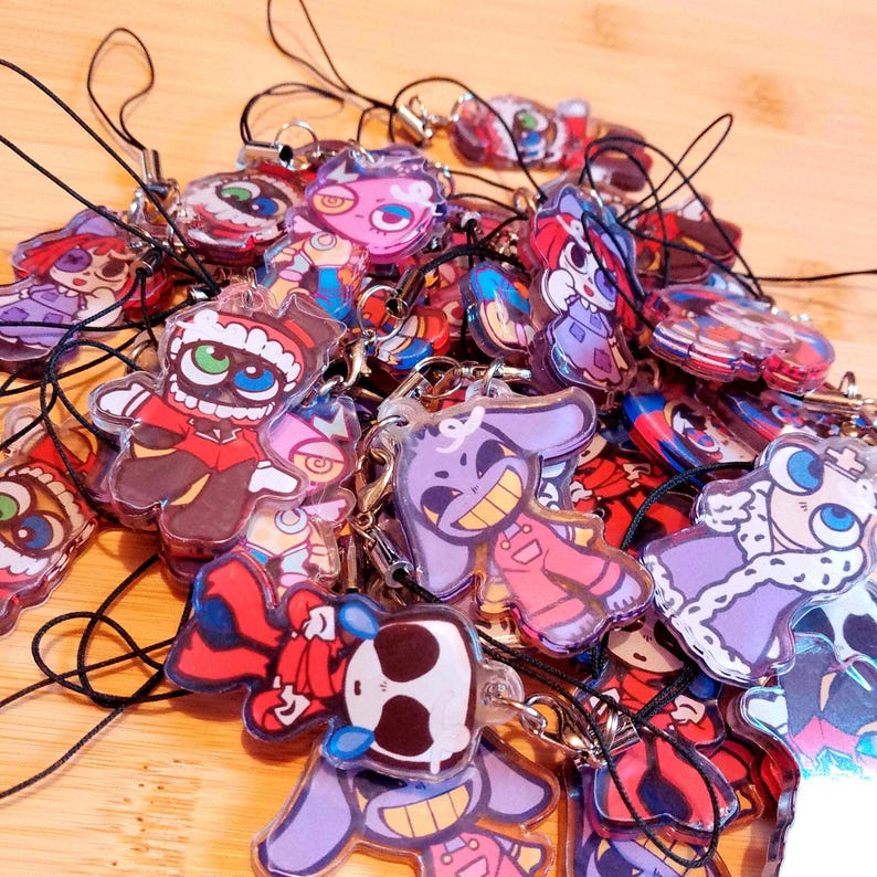 May include: A collection of colorful acrylic charms with cartoon character designs. The charms feature various figures in red, purple, and blue, each with a black string for attachment. The background is a light wood surface.
