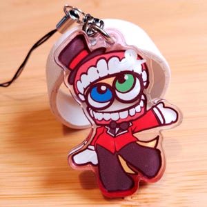 May include: A clear acrylic charm featuring a cartoon character in a red suit and top hat. The character has a wide grin, and eyes that are blue and green. The charm has a silver clasp and a black string.