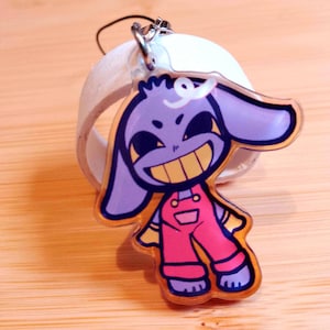 May include: A clear acrylic keychain featuring a cartoon character with purple skin, large ears, and a wide yellow grin. The character wears pink overalls with yellow accents. A silver-colored clasp and a white ring are attached.