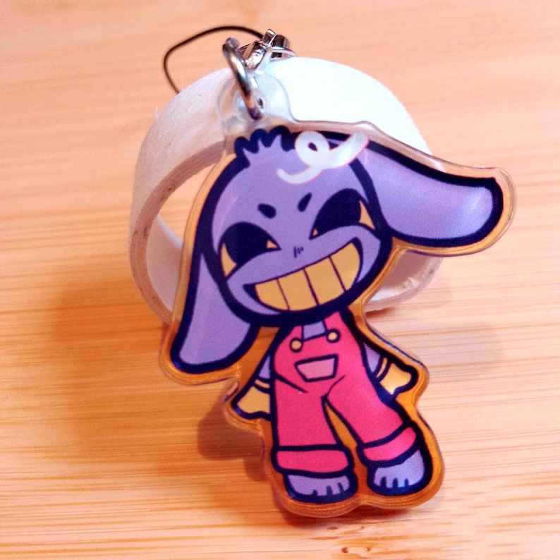 May include: A clear acrylic keychain featuring a cartoon character with purple skin, large ears, and a wide yellow grin. The character wears pink overalls with yellow accents. A silver-colored clasp and a white ring are attached.