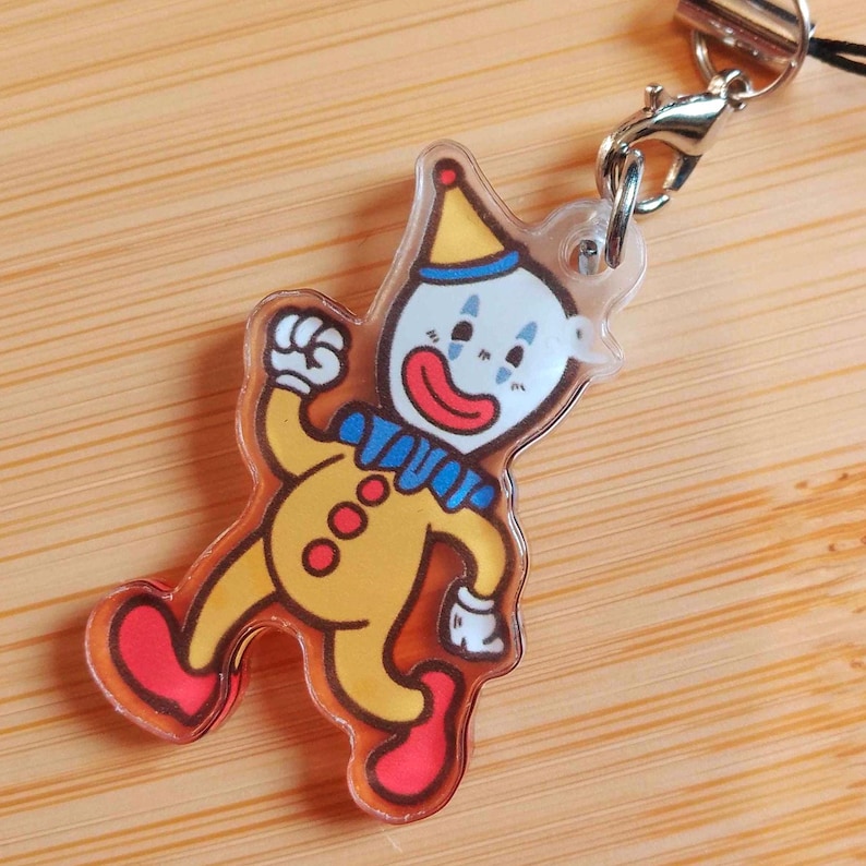 May include: A clear acrylic charm featuring a cartoon clown in yellow, red, and blue. The clown has a white face with red lips, blue teardrops, and a yellow hat. The charm is attached to a silver-colored clasp and black cord.
