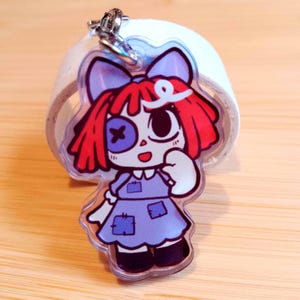 May include: A clear acrylic keychain featuring a cartoon doll with red hair, a blue bow, and a purple dress. The doll has one eye with a cross and a large eye. The keychain has a silver clasp.
