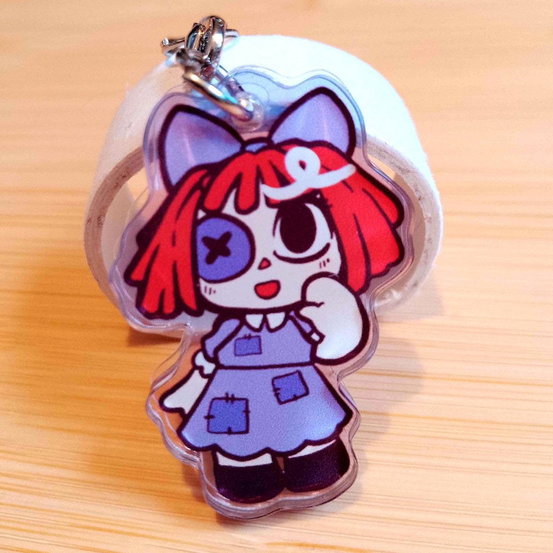 May include: A clear acrylic keychain featuring a cartoon doll with red hair, a blue bow, and a purple dress. The doll has one eye with a cross and a large eye. The keychain has a silver clasp.