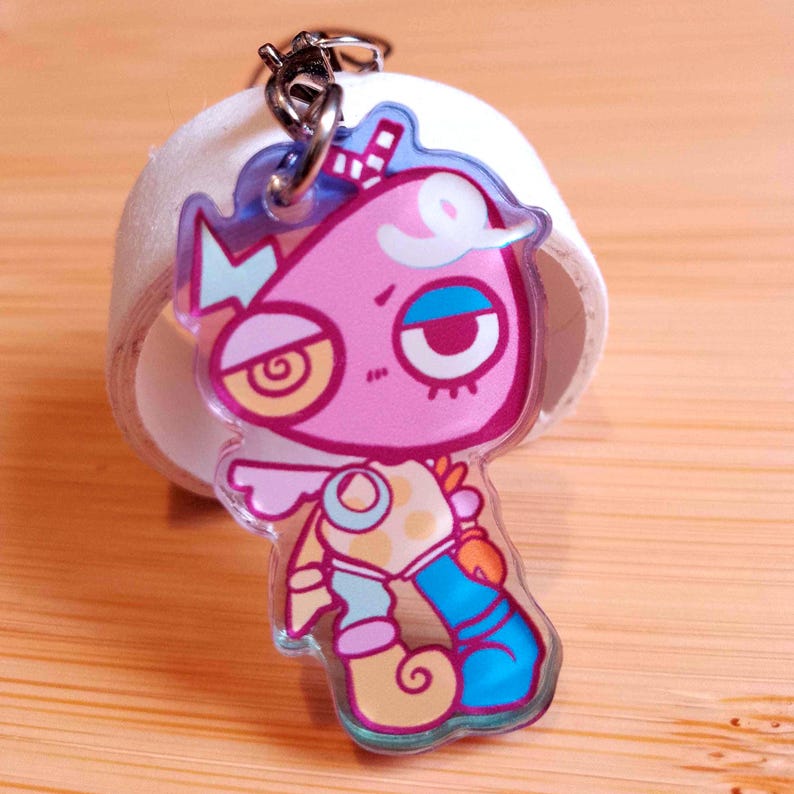 May include: A clear acrylic keychain featuring a cartoon character with a pink head, large yellow eye, and blue and yellow clothing. The keychain has a silver metal ring and is set against a light wooden surface.
