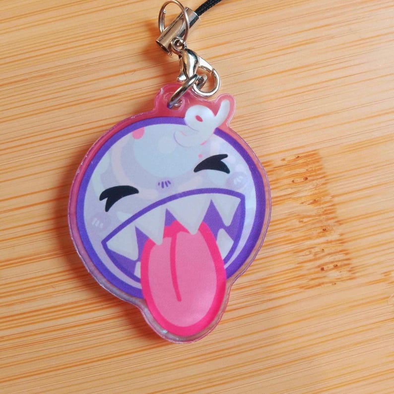 May include: A clear acrylic charm featuring a cartoon ghost with a pink tongue sticking out. The charm has a purple and pink border, black eyes, and a silver clasp with a black cord.