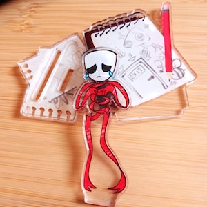 May include: A clear acrylic charm depicting a sad cartoon figure with red limbs and a white head with black eyes and a teardrop. A notepad with drawings and a red pencil are behind the figure. The charm is on a wooden surface.