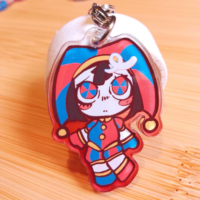 May include: A clear acrylic keychain featuring a cartoon jester character with a red, blue, and yellow color scheme. The jester has a sad expression and a matching hat. The keychain has a silver metal clasp.