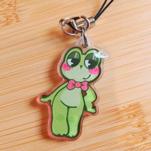May include: A clear acrylic charm featuring a cartoon frog with a pink bow tie and rosy cheeks. The frog is green with dark green accents and a red outline. It has a silver clasp and a black cord for attachment. The charm is approximately 2 inches in height.