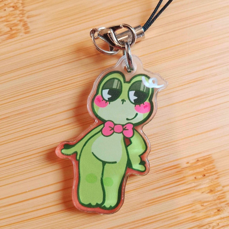 May include: A clear acrylic charm featuring a cartoon frog with a pink bow tie and rosy cheeks. The frog is green with dark green accents and a red outline. It has a silver clasp and a black cord for attachment. The charm is approximately 2 inches in height.