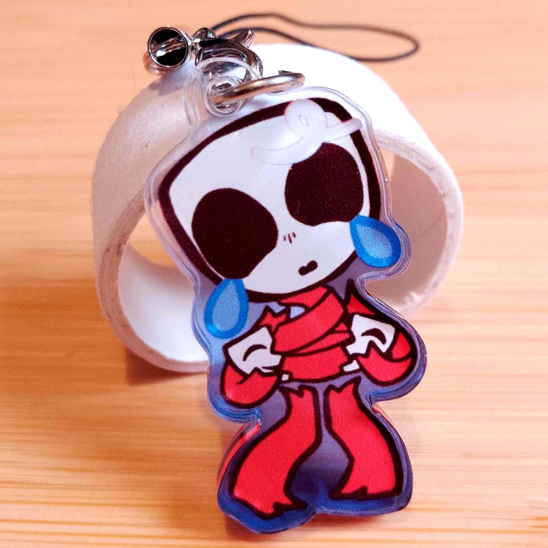 May include: A clear acrylic charm featuring a cartoon character with a white head, large black eye sockets, and red clothing. The character has blue teardrops and is attached to a silver keychain.