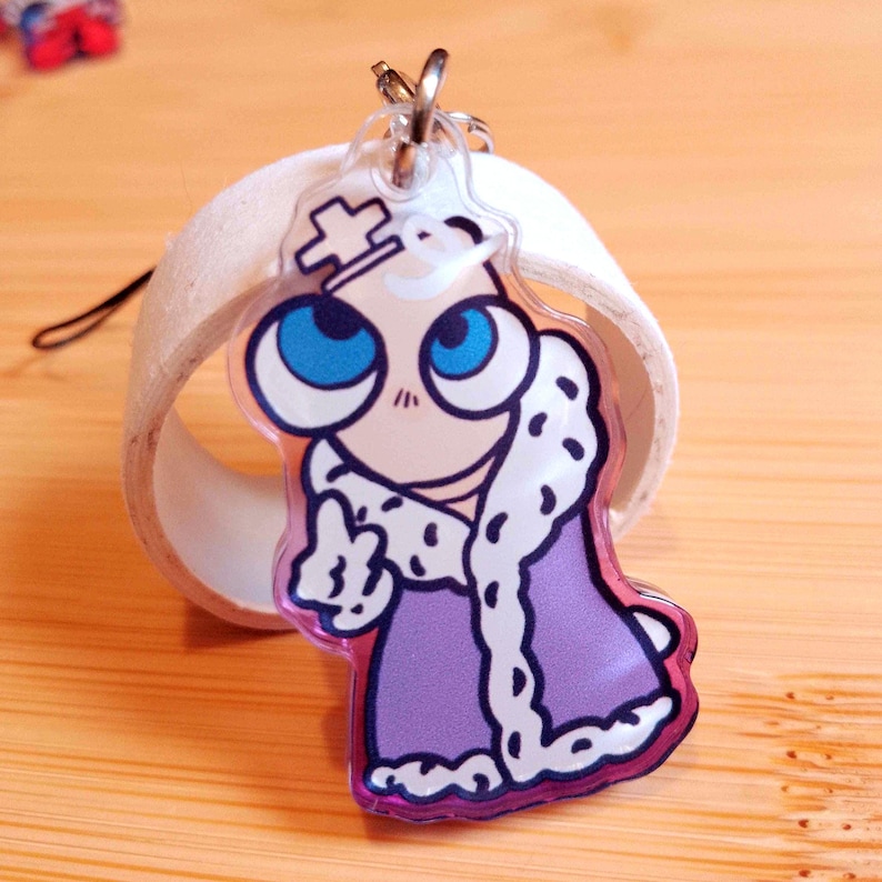 May include: A clear acrylic keychain featuring a cartoon character with large blue eyes, a crown, and a purple robe with white trim. The keychain has a silver ring and a black string attached.