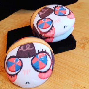 May include: Two plush character pins, round in shape, with large eyes. The pins have a white base with brown, pink, blue, and orange details. They are placed on a black surface, against a wooden background.