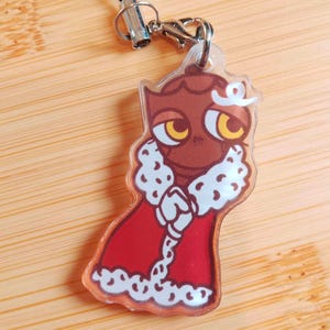 May include: A clear acrylic charm featuring a cartoon owl character wearing a red robe with white fur trim. The owl has brown feathers, large yellow eyes, and a white crown. The charm is attached to a silver-colored clasp and black lanyard.