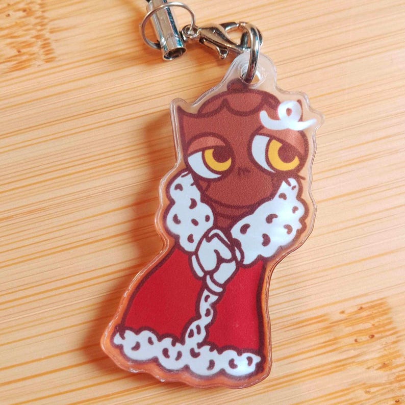 May include: A clear acrylic charm featuring a cartoon owl character wearing a red robe with white fur trim. The owl has brown feathers, large yellow eyes, and a white crown. The charm is attached to a silver-colored clasp and black lanyard.