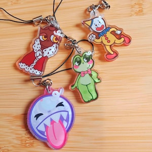 May include: Four colorful acrylic charms with black cords. The charms feature cartoon designs: a red owl, a yellow clown, a green frog with a pink bow tie, and a purple ghost with a pink tongue. Each charm has a silver-colored clasp.