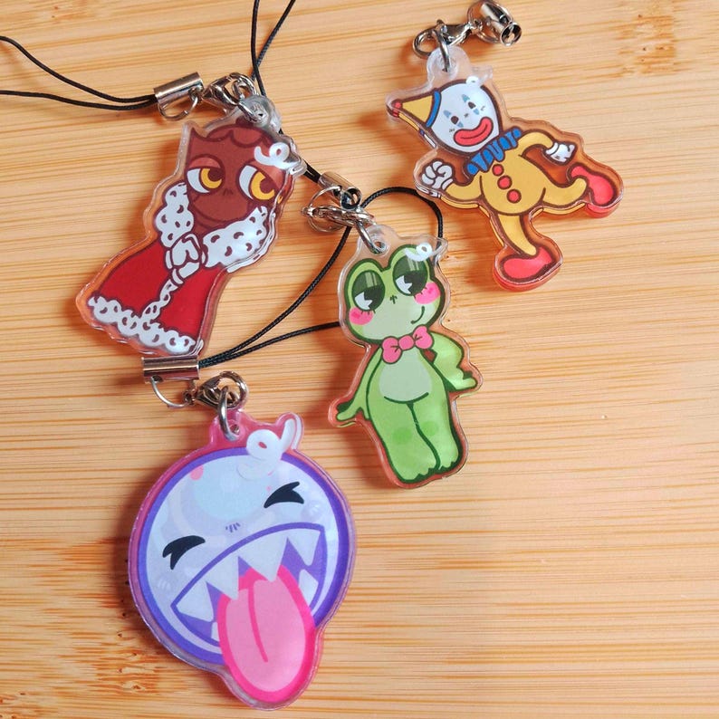 May include: Four colorful acrylic charms with black cords. The charms feature cartoon designs: a red owl, a yellow clown, a green frog with a pink bow tie, and a purple ghost with a pink tongue. Each charm has a silver-colored clasp.