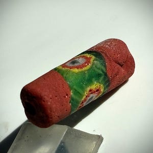 Pode incluir: Uma conta cilíndrica vermelha com um padrão decorativo verde e amarelo. A conta tem uma superfície texturizada e um buraco que atravessa o seu centro. O design inclui formas semelhantes a olhos.