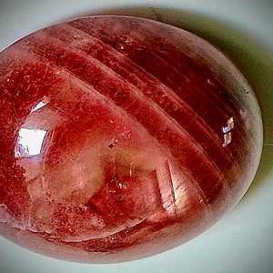 May include: A polished, oval-shaped gemstone with a translucent, reddish-brown hue. The stone features subtle, lighter-colored bands that run across its surface. The gem reflects light, creating highlights and shadows.
