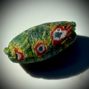 Pode incluir: Uma conta de vidro oblonga com uma base verde, detalhes amarelos e vermelhos e padrões circulares brancos e azuis. A conta tem uma superfície texturizada e uma aparência única, artesanal. A conta é provavelmente usada para fazer joias ou para fins decorativos.