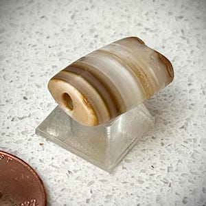 May include: A banded agate bead with a hole drilled through the centre. The bead is a light brown colour with white and brown stripes.