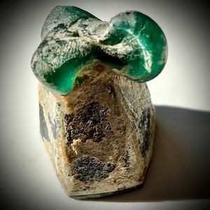 May include: A unique, abstract sculpture featuring a vibrant green, rounded top section atop a textured, beige base. The base has dark, mottled patterns and a rough, porous surface, creating a striking contrast with the smooth, translucent green top.