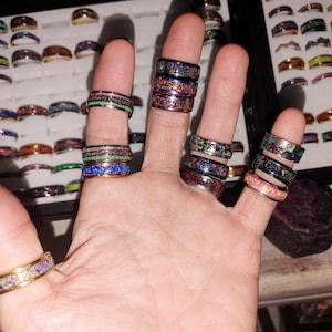 May include: A hand displaying multiple rings with colorful, patterned designs. The rings feature various colors, including green, blue, and pink, and are displayed against a backdrop of other rings in a display case. The rings are a mix of styles and sizes.