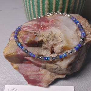 Puede incluir: Una delicada pulsera de cuentas con piedras azules y grises, colocada sobre una superficie de piedra natural. La pulsera presenta una cadena y un cierre plateados. La imagen también incluye una maceta de cerámica a rayas verdes y blancas y una tarjeta de visita con el texto "Mags Bags Crafts".