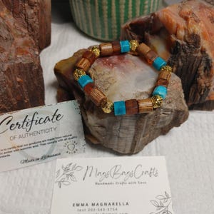 May include: A handmade bracelet featuring wooden, turquoise, and gold-colored beads. The bracelet is displayed on a rock with a certificate of authenticity and a business card from Mags Bags Crafts.