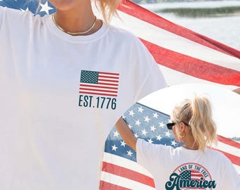 May include: White t-shirt with a small American flag graphic and the text "EST. 1776" on the front. The back of the shirt features a circular design with the words "Land of the Free America EST. 1776 Because of the Brave" and another American flag graphic.