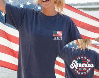 May include: Navy blue t-shirt with a small American flag graphic and "EST. 1776" on the front. The back of the shirt features a circular design with "America" and "Land of the Free Because of the Brave".