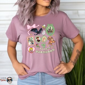 May include: Mauve t-shirt featuring a collection of cartoon illustrations, including a dragon, castle, and characters from various franchises. The word "UNIVERSE" is spelled out in a string of beads. A pink bow is at the top.