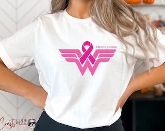 May include: White t-shirt with a pink Wonder Woman logo and a pink ribbon. The text '#team emma' is printed on the shirt.