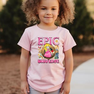 May include: A light pink t-shirt featuring the words "EPIC UNIVERSE" in pink block letters. The shirt has a graphic of a cartoon princess and other characters. The shirt is being worn by a young child.