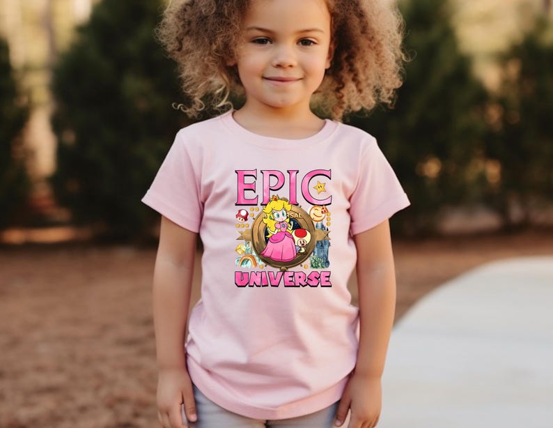 May include: A light pink t-shirt featuring the words "EPIC UNIVERSE" in pink block letters. The shirt has a graphic of a cartoon princess and other characters. The shirt is being worn by a young child.