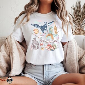 May include: White t-shirt featuring a colorful graphic design with the text "Epic Universe". The design includes cartoon characters and elements from various franchises, such as a dragon, a castle, and video game characters.