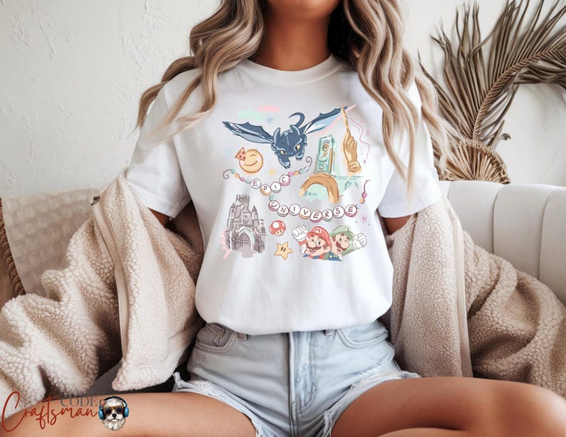 May include: White t-shirt featuring a colorful graphic design with the text "Epic Universe". The design includes cartoon characters and elements from various franchises, such as a dragon, a castle, and video game characters.