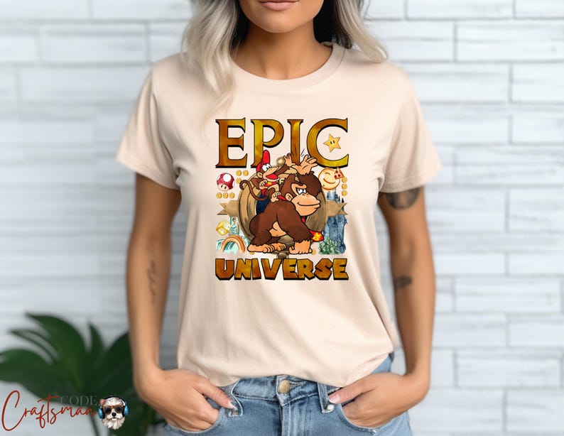 May include: Beige t-shirt featuring a colorful graphic with the text "EPIC UNIVERSE". The graphic includes cartoon characters, a star, and a mushroom. The shirt is a crew neck and has short sleeves.