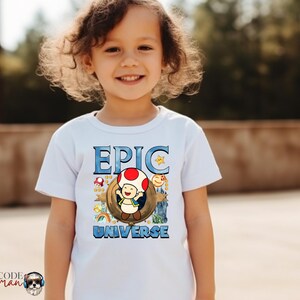 May include: A white t-shirt featuring the words "EPIC UNIVERSE" in blue, with a cartoon character design. The design includes a mushroom, a star, and other colorful elements. The shirt is worn by a young child.