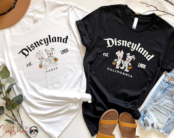 May include: Two white t-shirts with a black and white graphic of Mickey Mouse and Minnie Mouse dressed as mummies. The text on the shirts reads "Disneyland est. 1955" with "Paris" on one shirt and "California" on the other.