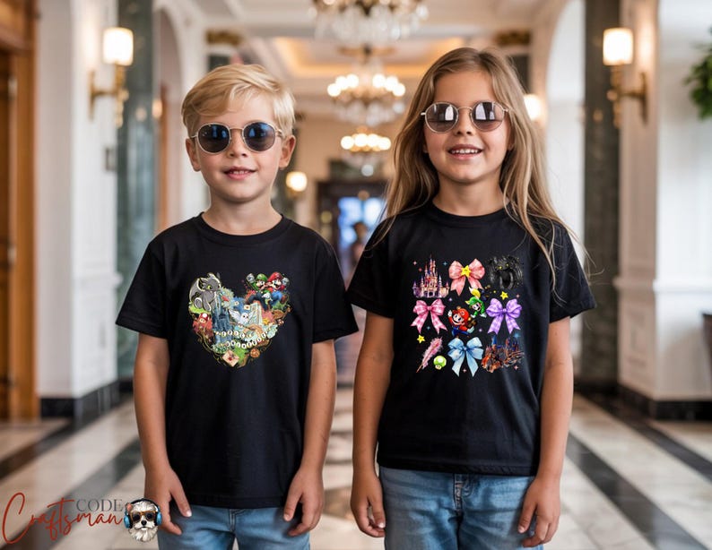 May include: Two children wearing black t-shirts and sunglasses. The t-shirts feature colorful graphic designs. One shirt has a heart-shaped design with cartoon characters. The other shirt has a design with bows and a castle.