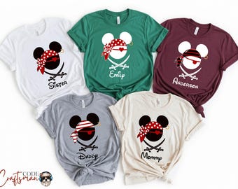 May include: Five short-sleeved t-shirts in white, green, burgundy, gray, and cream. Each shirt features a pirate-themed Mickey Mouse design with a red and white bandana, crossed swords, and text such as "Sister", "Emily", "Anderson", "Daddy", and "Mommy".