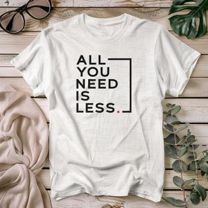 May include: White t-shirt with the text "ALL YOU NEED IS LESS" in black, enclosed in a black rectangle. The word "LESS" has a red period at the end. Black sunglasses and a beige cloth are in the background.