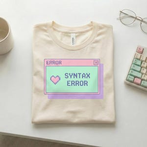 Könnte beinhalten: Cremefarbenes T-Shirt mit einer pixeligen "ERROR"-Nachricht und dem Text "SYNTAX ERROR" in einem Fenster im Retro-Computer-Stil. Eine Pixel-Herz-Grafik ist enthalten. Das Shirt ist sauber auf einer weißen Oberfläche gefaltet, mit Tastatur und Brille.