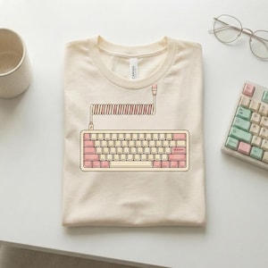 Puede incluir: Camiseta color crema con un gráfico de un teclado rosa y beige con un cable en espiral. El diseño del teclado incluye teclas rosas y un marco beige. Vista parcial de un teclado y gafas en el fondo.