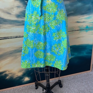 Può includere: Gonna a trapezio con sfondo blu vibrante e motivo floreale verde. La gonna ha una cintura abbinata che si annoda in vita. La gonna è esposta su un manichino vintage.