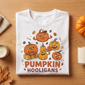 May include: A white t-shirt featuring a graphic print of five cartoon pumpkins with various facial expressions. The text PUMPKIN HOOLIGANS is printed in orange and brown below the pumpkins, surrounded by small autumn leaves, acorns, and twigs.